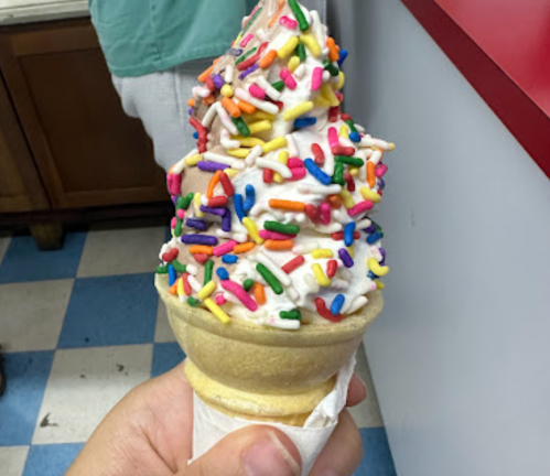A hand holding a soft-serve ice cream cone topped with colorful sprinkles.