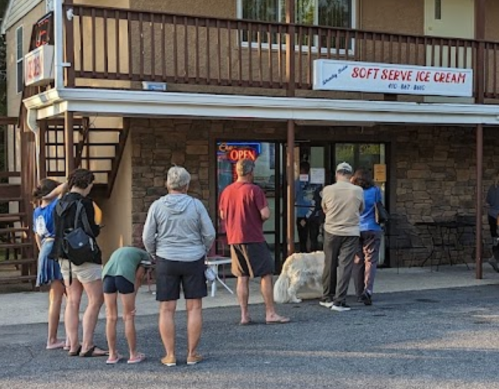 A line of people waiting outside a soft serve ice cream shop, with a dog nearby and a sign indicating it's open.