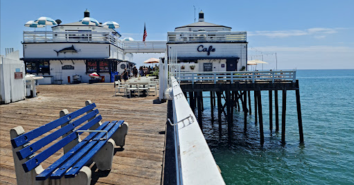 A seaside pier with a cafe, blue benches, and umbrellas, overlooking calm waters under a clear sky.