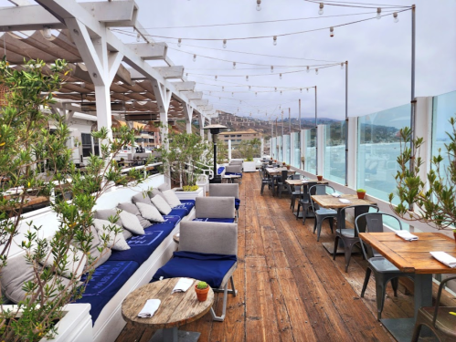 A stylish outdoor restaurant patio with wooden flooring, cozy seating, and string lights, overlooking a scenic view.