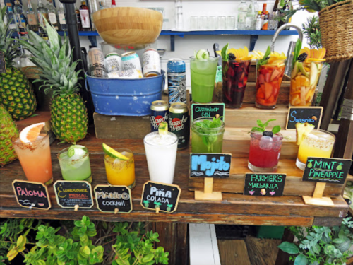 A colorful display of various cocktails and drinks on a wooden bar, surrounded by fruits and greenery.