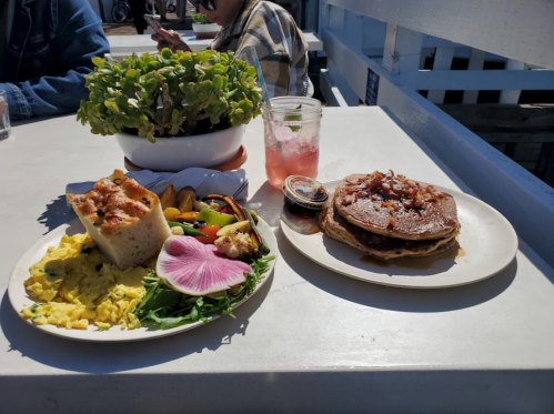 A plate of scrambled eggs, vegetables, and bread alongside pancakes topped with bacon, with a drink and a plant in the background.