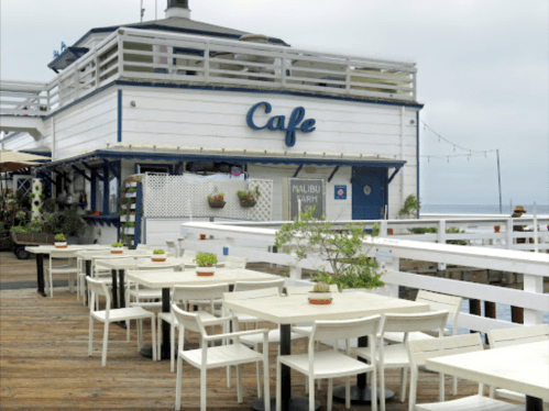 A seaside café with white tables and chairs, featuring potted plants, against a cloudy sky.