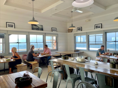 Bright, airy café with wooden tables, diners enjoying meals, and large windows overlooking the ocean.