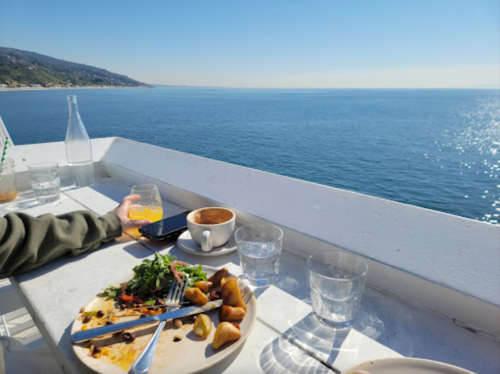 A table by the ocean with a half-eaten meal, a coffee cup, and a glass, overlooking a clear blue sea.
