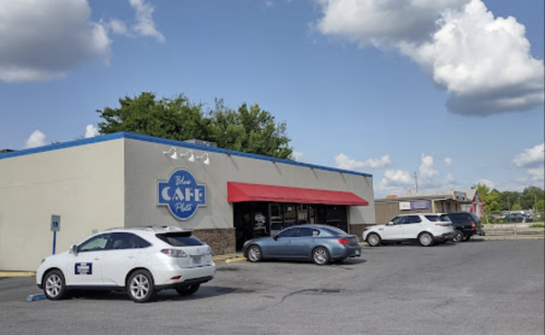 A café building with a red awning, parked cars, and a blue sky with fluffy clouds.