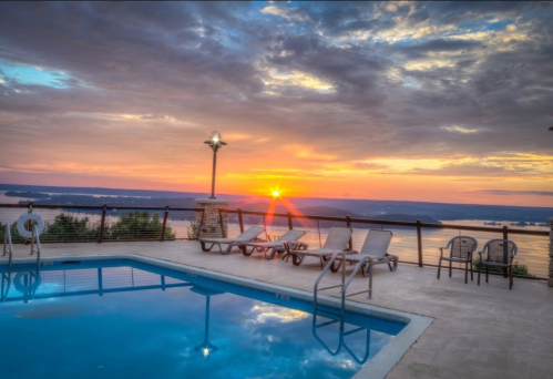 Sunset over a pool with lounge chairs, surrounded by scenic hills and a calm lake, under a cloudy sky.