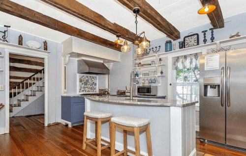 Cozy kitchen with wooden beams, granite island, blue cabinetry, and stainless steel appliances, featuring a staircase in the background.