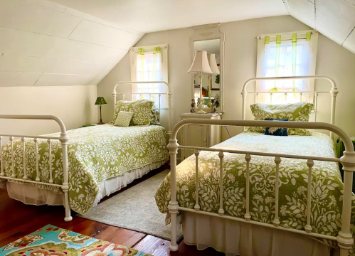 Cozy attic bedroom with two white metal beds, green floral bedding, and a mirror, featuring soft natural light.