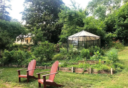 A serene garden with two red chairs, lush greenery, and a glass greenhouse in the background.