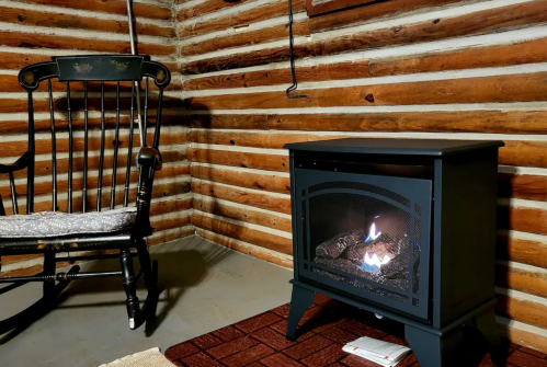 A cozy cabin interior featuring a black electric fireplace and a wooden rocking chair against log walls.