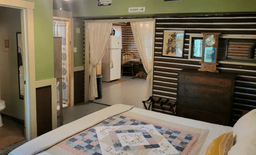 Cozy bedroom with a quilt, leading to a rustic kitchen and dining area through an open doorway.