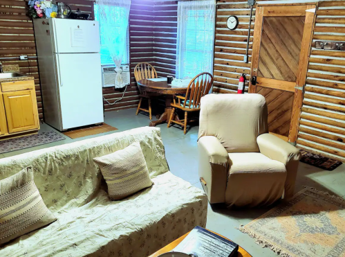 Cozy cabin interior with a sofa, armchair, dining table, and kitchenette, featuring wooden walls and rustic decor.