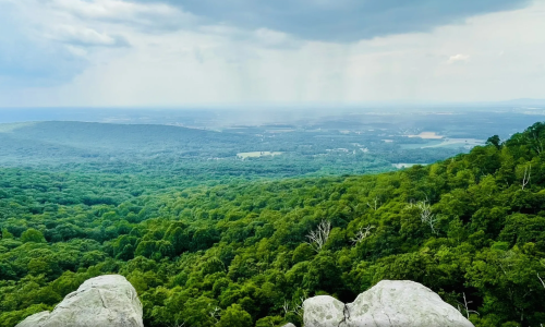 A panoramic view of lush green mountains and valleys under a cloudy sky, with rocky foreground.
