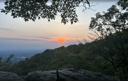 A sunset over a mountainous landscape, framed by trees and rocky outcrops.