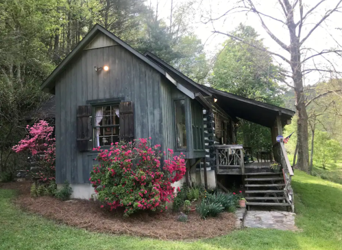 A charming wooden cabin surrounded by greenery and vibrant pink flowers, with a porch and steps leading to the entrance.
