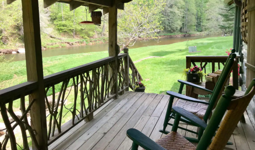A peaceful porch view overlooking a river and lush greenery, featuring rocking chairs and potted flowers.