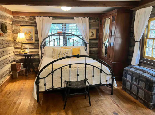 Cozy bedroom with a wooden log wall, iron bed, white linens, and a vintage chest, illuminated by soft natural light.