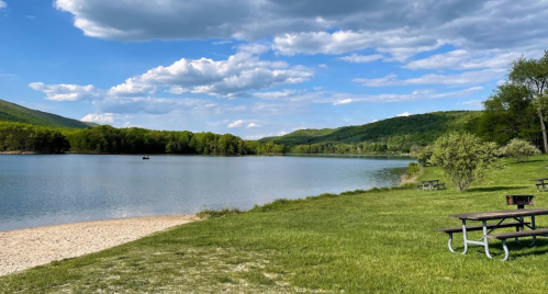 A serene lake surrounded by green hills, with picnic tables on the grassy shore and fluffy clouds in a blue sky.