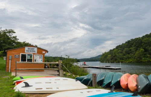A boat rental shop by a calm lake, with kayaks lined up on the grass and a dock in the background under cloudy skies.