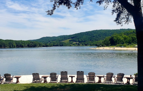 A serene lake view with wooden chairs lined up on the shore, surrounded by green hills and a clear blue sky.