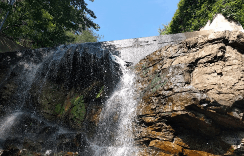 A waterfall cascading over rocky terrain, surrounded by lush greenery and a clear blue sky above.