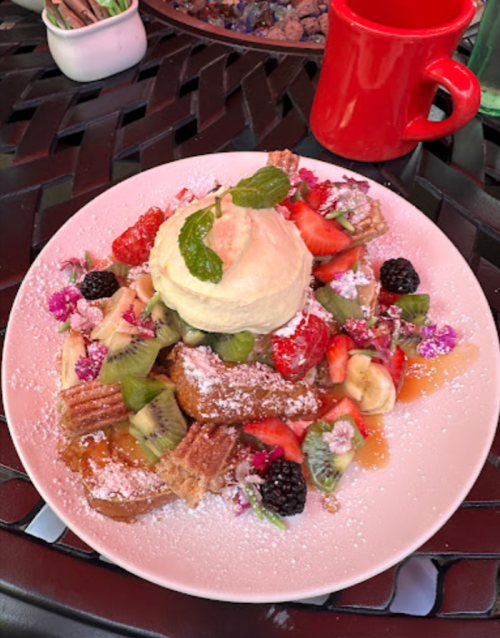 A colorful plate of fruit, waffles, and ice cream, garnished with mint and edible flowers, on a black woven table.