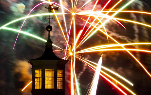 Colorful fireworks burst in the night sky above a silhouetted tower with a weather vane.