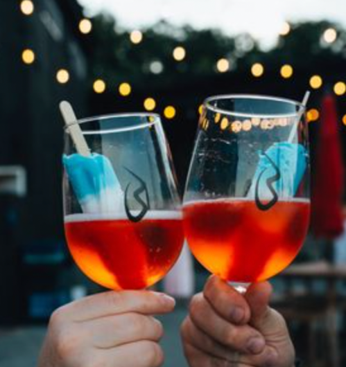 Two glasses of vibrant red drinks with blue popsicles, clinking together against a backdrop of string lights.