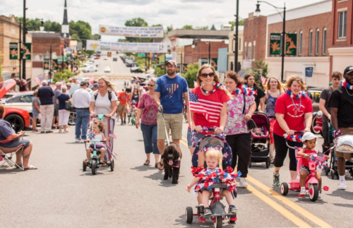 A festive parade scene with families, children on tricycles, and a dog, celebrating with American flags and decorations.