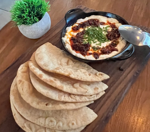 A wooden platter with flatbreads and a small skillet of creamy dip topped with herbs and sauce, alongside a decorative plant.
