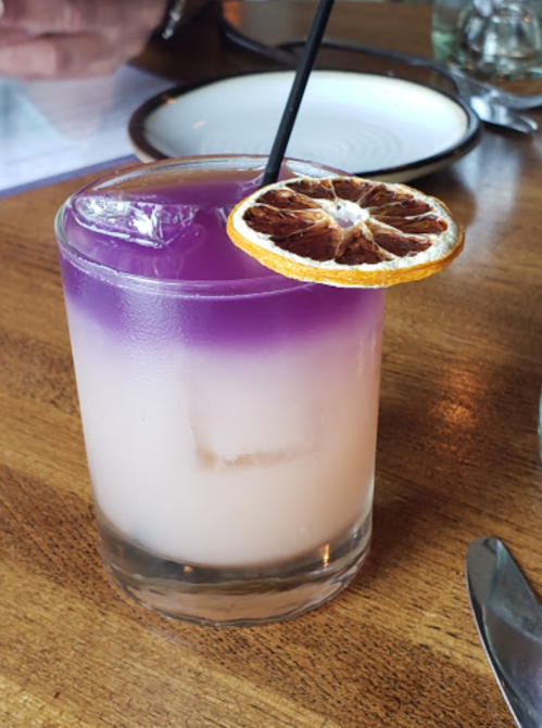 A layered cocktail in a glass, featuring purple and white hues, garnished with a dried orange slice.