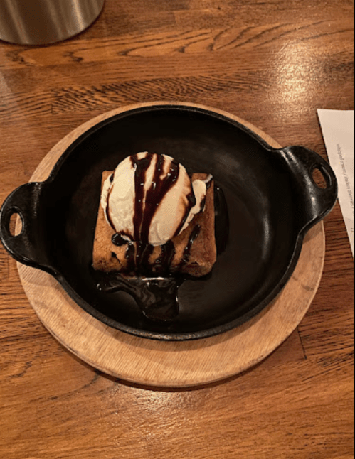A warm dessert in a cast-iron skillet, featuring a brownie topped with vanilla ice cream and chocolate sauce.