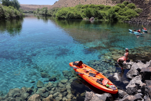 A serene river scene with clear blue water, kayaks, and a person sitting on rocks by the shore.