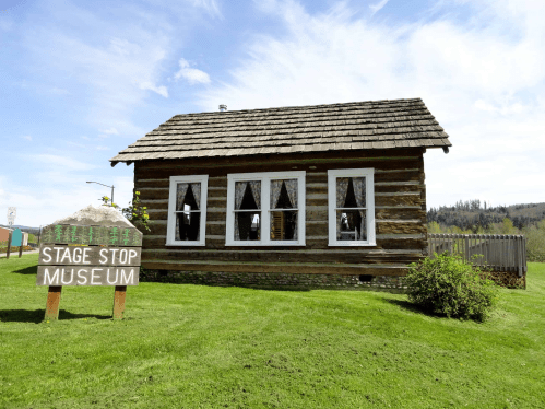 A rustic log cabin with a sign reading "Stage Stop Museum" in front, surrounded by green grass and trees.