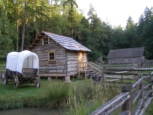 A rustic log cabin and a covered wagon in a forested area, surrounded by greenery and wooden fences.