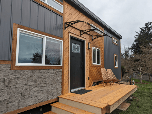 A modern tiny house with a wooden deck, large windows, and a mix of wood and stone exterior finishes.