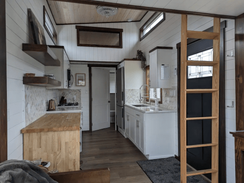 A cozy, modern tiny kitchen with wooden accents, white cabinets, and a ladder leading to a loft area.