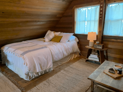 Cozy bedroom with a wooden interior, featuring a bed, bedside lamp, and a small table with decor. Natural light from windows.