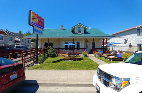 A restaurant with a green roof and outdoor seating, surrounded by grass and parked cars on a sunny day.