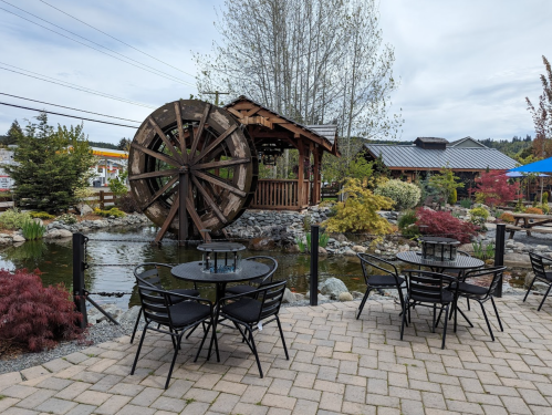A scenic outdoor area with a water wheel, pond, and tables surrounded by greenery and colorful plants.