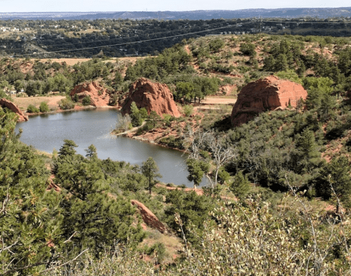 Scenic view of red rock formations beside a calm lake, surrounded by green trees and rolling hills in the background.