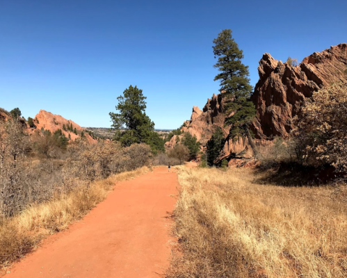 A dirt path winds through a rocky landscape with sparse trees and dry grass under a clear blue sky.