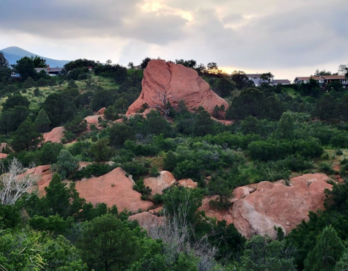 A large red rock formation surrounded by green trees and shrubs under a cloudy sky.
