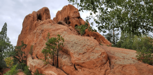 A rugged red rock formation surrounded by greenery under a cloudy sky.