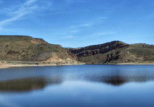 A serene lake surrounded by hills under a clear blue sky, reflecting the landscape in calm waters.