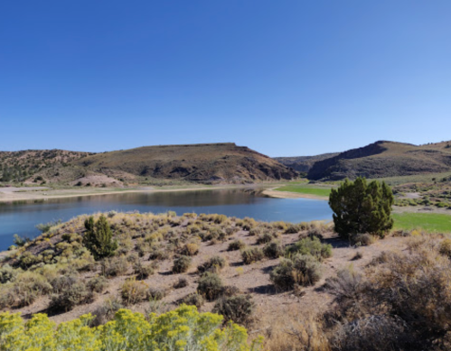 A serene landscape featuring a calm lake surrounded by hills and sparse vegetation under a clear blue sky.