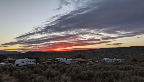 Sunset over a desert landscape with RVs and a shelter, surrounded by sagebrush and distant mountains.