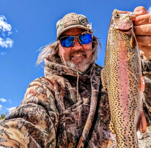 A smiling person in a camo hoodie holds a colorful trout against a clear blue sky.