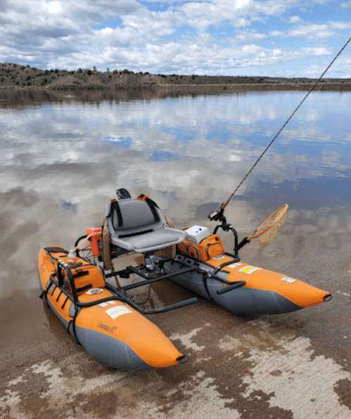 An orange inflatable kayak with a seat and fishing gear, anchored by the shore of a calm lake under a cloudy sky.